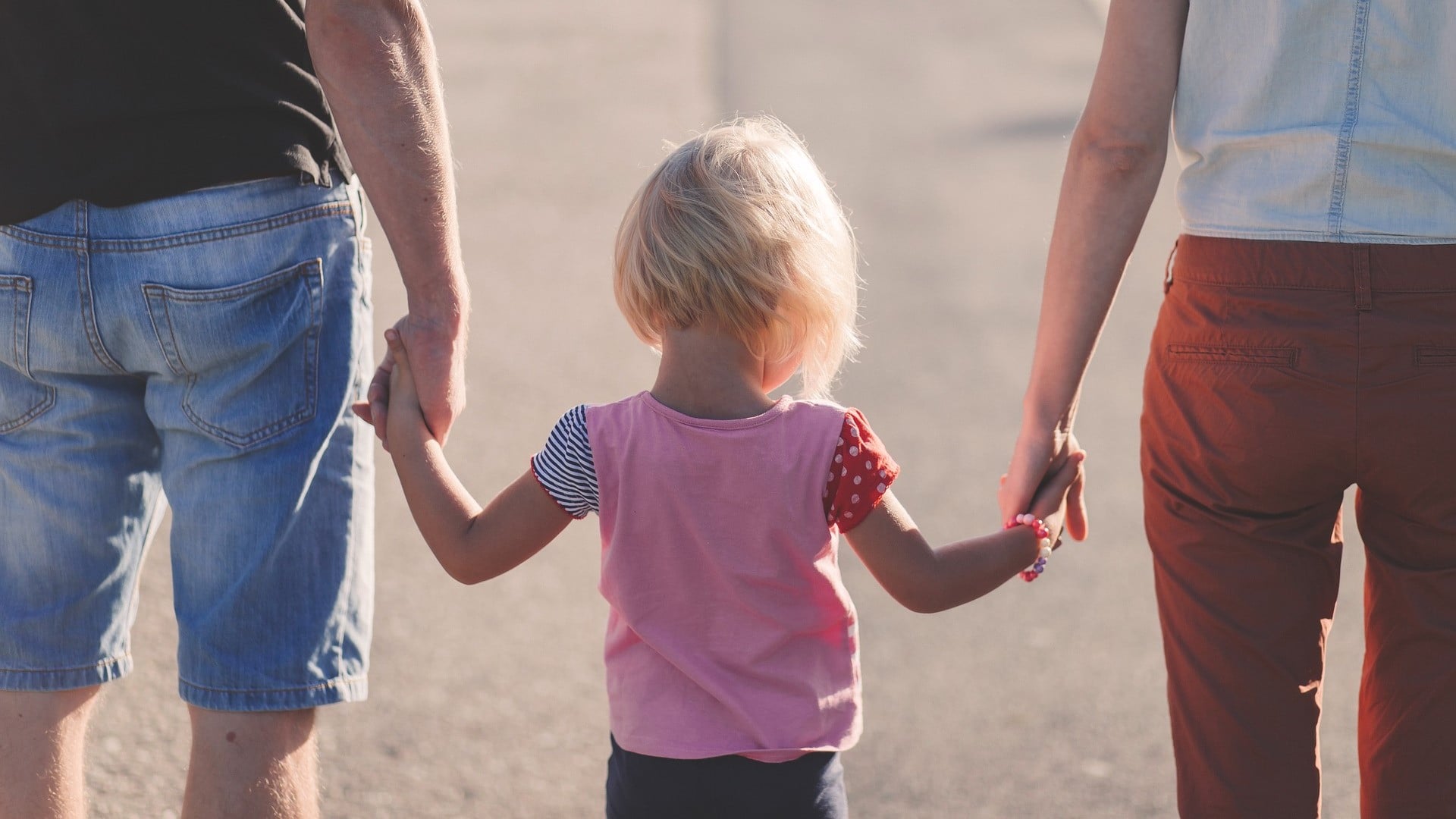 Padres caminando de espaldas con su hija pequeña, agarrándola de las manos.